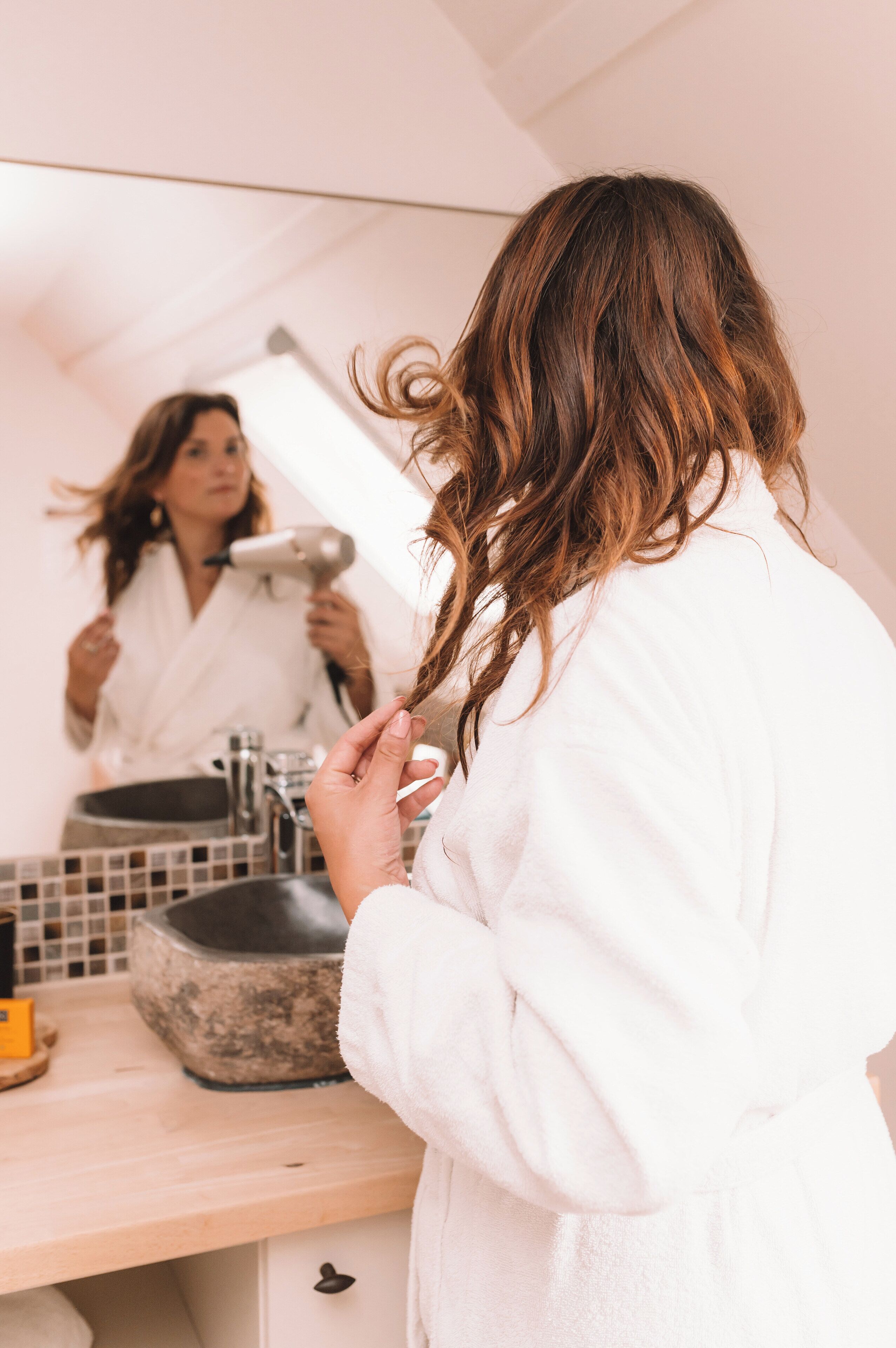 Een persoon in een witte badjas voor een spiegel in de badkamer, met een haardroger in de hand.