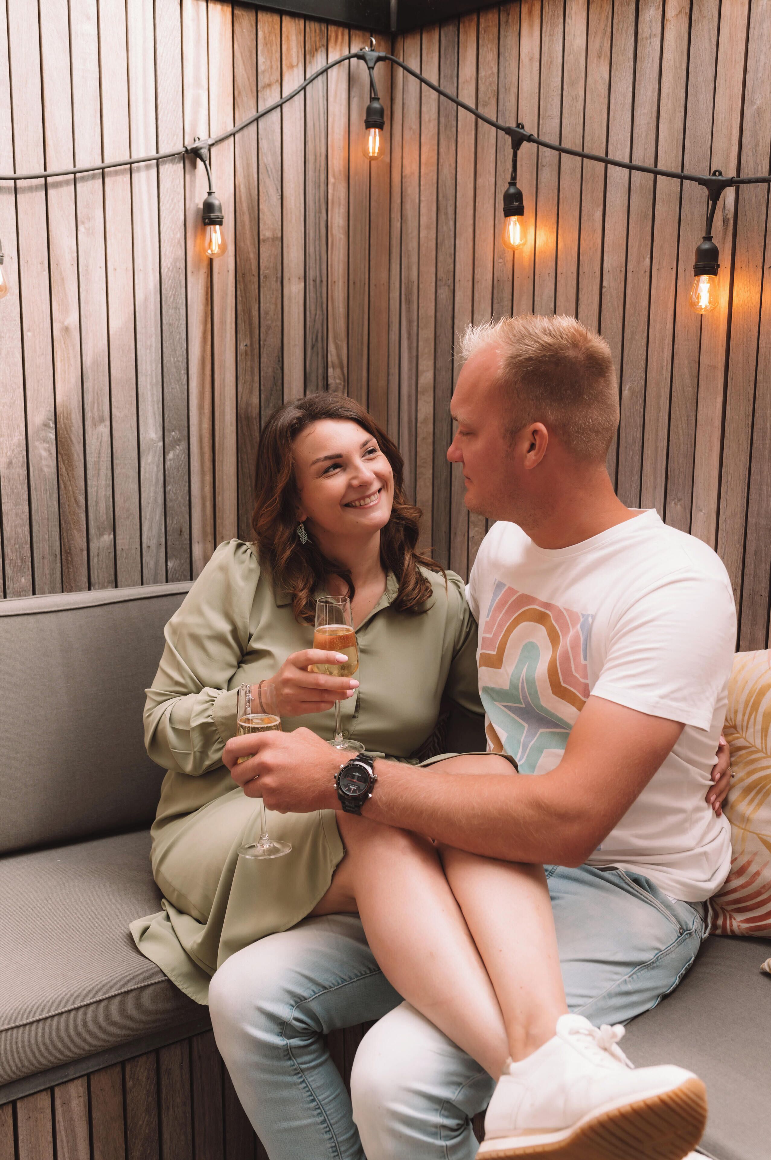 A couple sitting on a wooden bench with string lights above them.