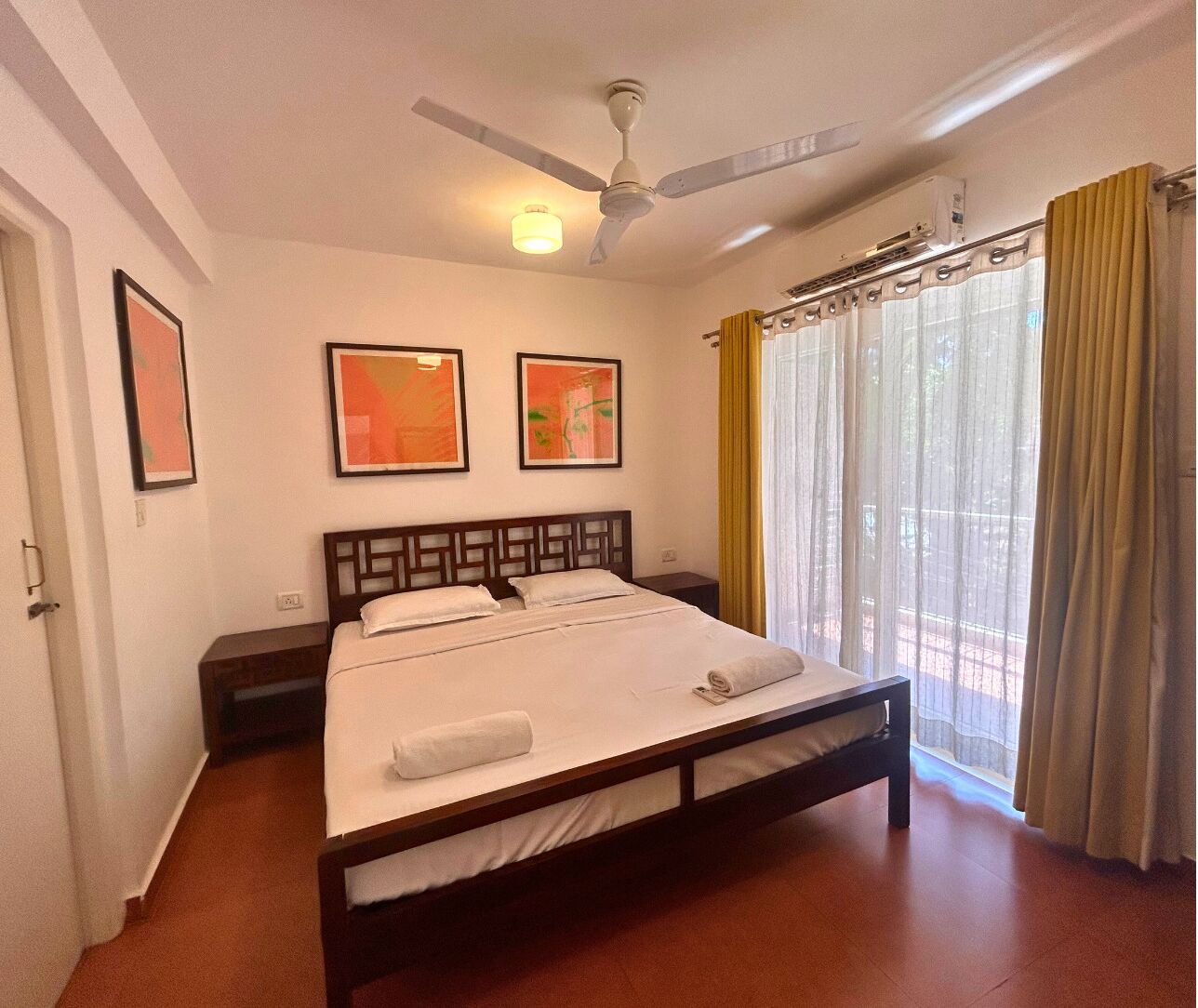 A bedroom with a bed, a ceiling fan, two framed pictures, a window with curtains, and a nightstand.