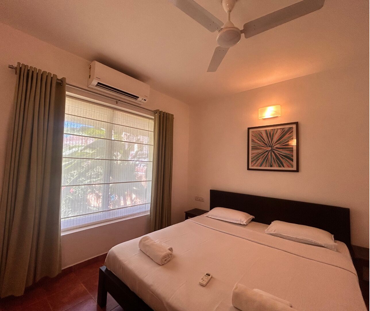 Una camera d'albergo con un letto, una ventola a soffitto, un condizionatore d'aria, una finestra con tende e un lampadario a parete.