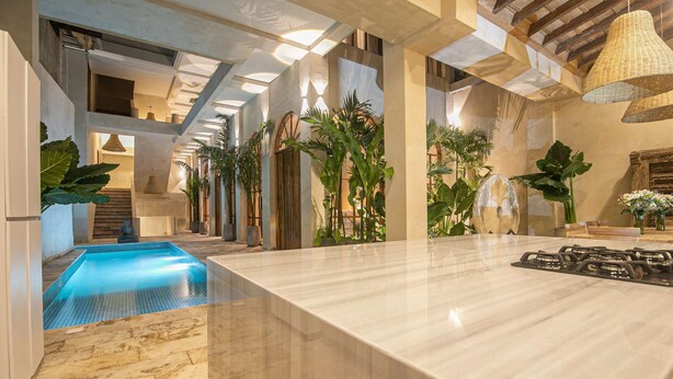 Un área de piscina interior moderna con suelo de mármol, rodeada de plantas y una escalera de madera.