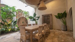 Un comedor con una larga mesa de madera y sillas de mimbre, rodeado de abundante follaje y una rústica puerta de madera.