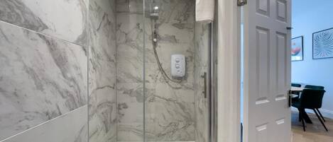 A glass-enclosed shower with marble walls, a white floor, and a door leading to a room with a dining area.