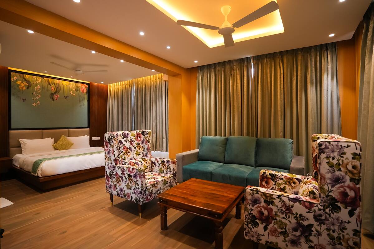 A hotel room with a bed, two floral armchairs, a sofa, a wooden coffee table, and a ceiling fan.
