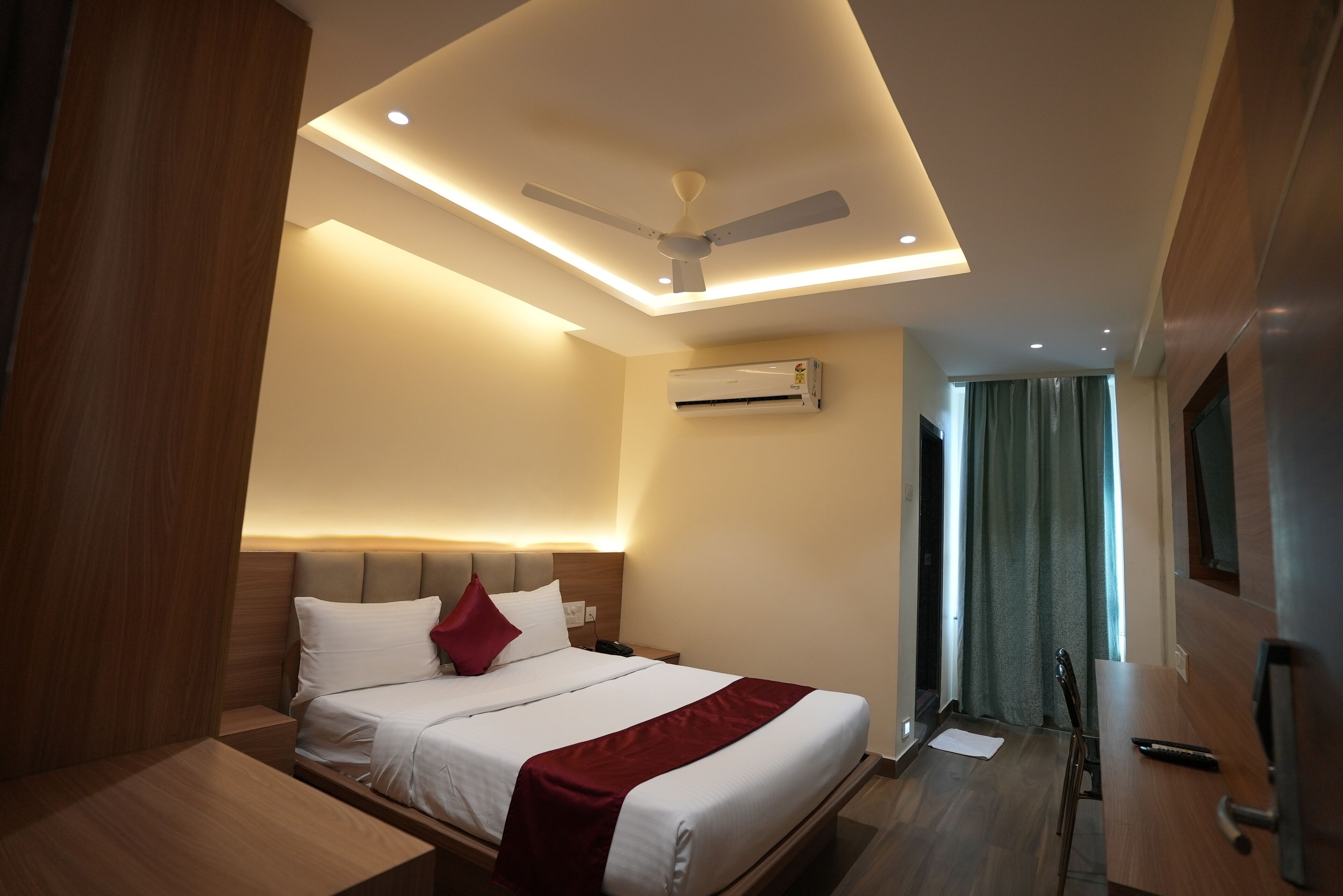A modern hotel room with a bed, a desk, a ceiling fan, recessed lighting, and an air conditioning unit.