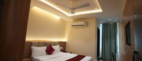 A modern hotel room with a bed, a desk, a ceiling fan, recessed lighting, and an air conditioning unit.