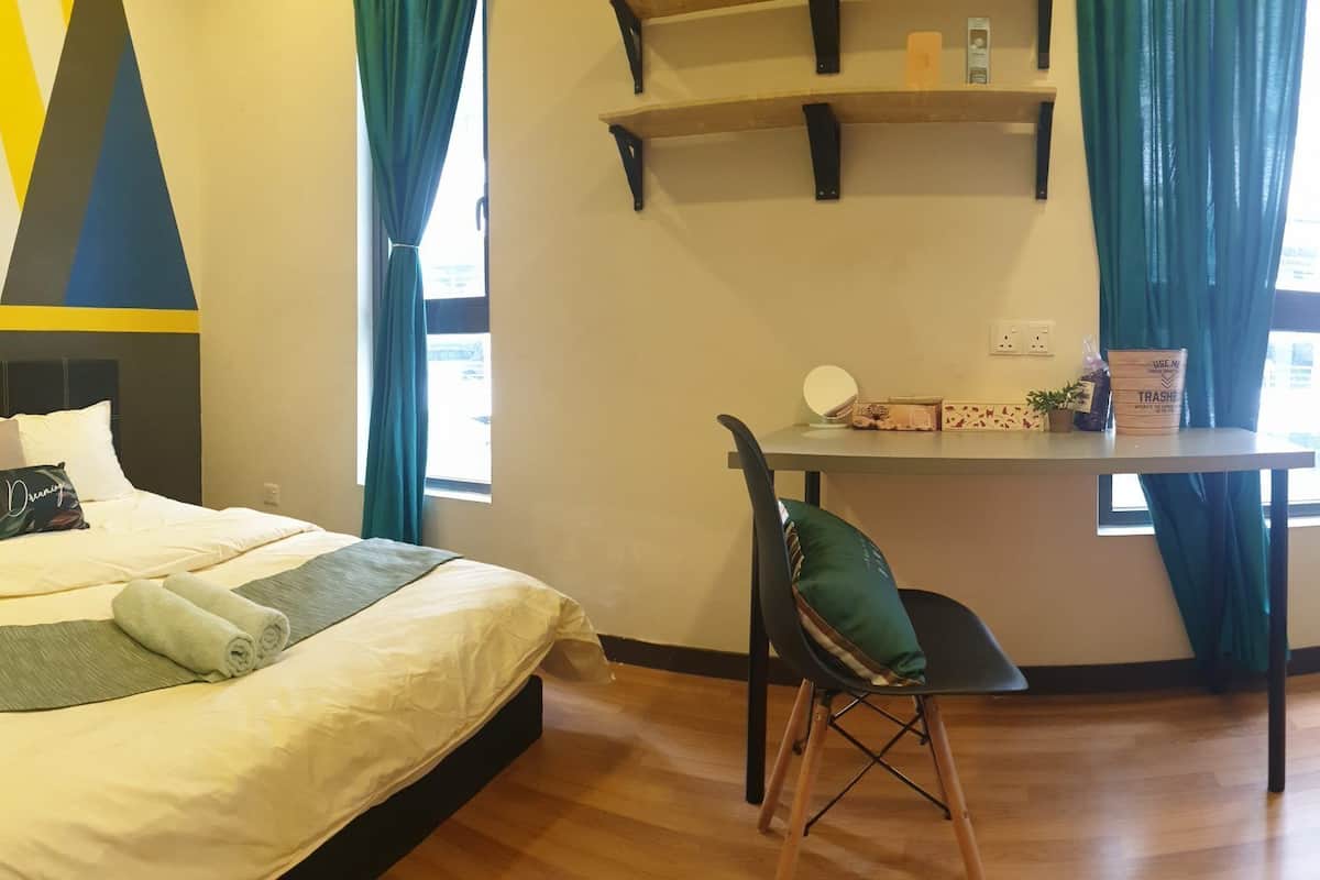 A hotel room with a bed, a desk, and a chair. There is a window with curtains, a wall with geometric patterns, and a shelf with toiletries.