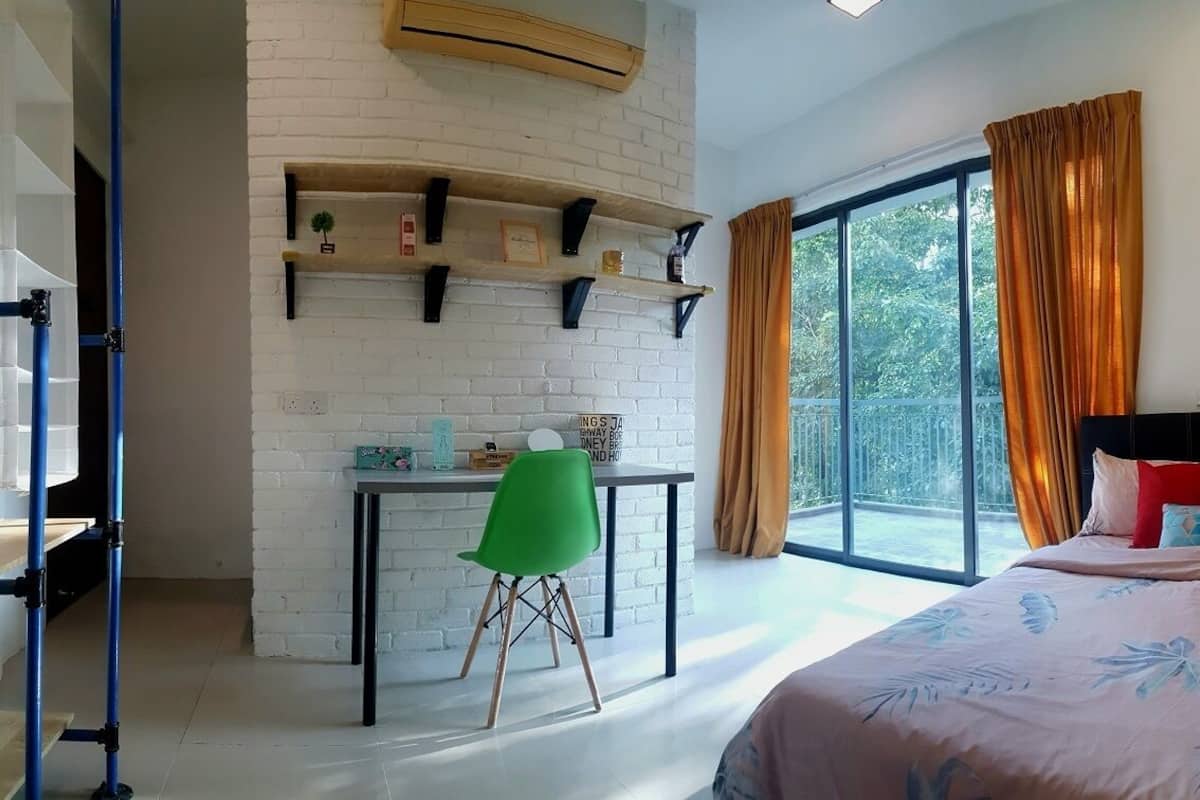 A bedroom with a bed, a desk with a chair, a window with curtains, and a brick wall with shelves.