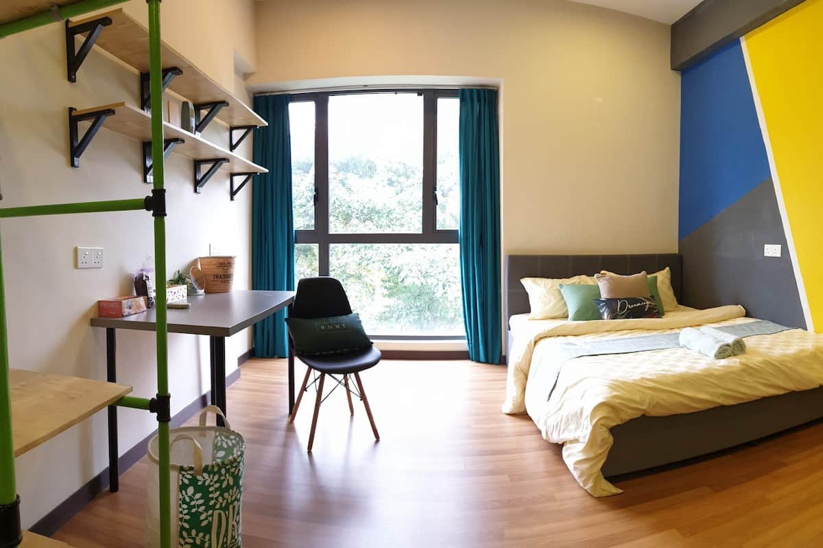 A modern hotel room with a bed, a desk, and a chair. There is a window with curtains, a view of greenery outside, and a wall with a geometric design.