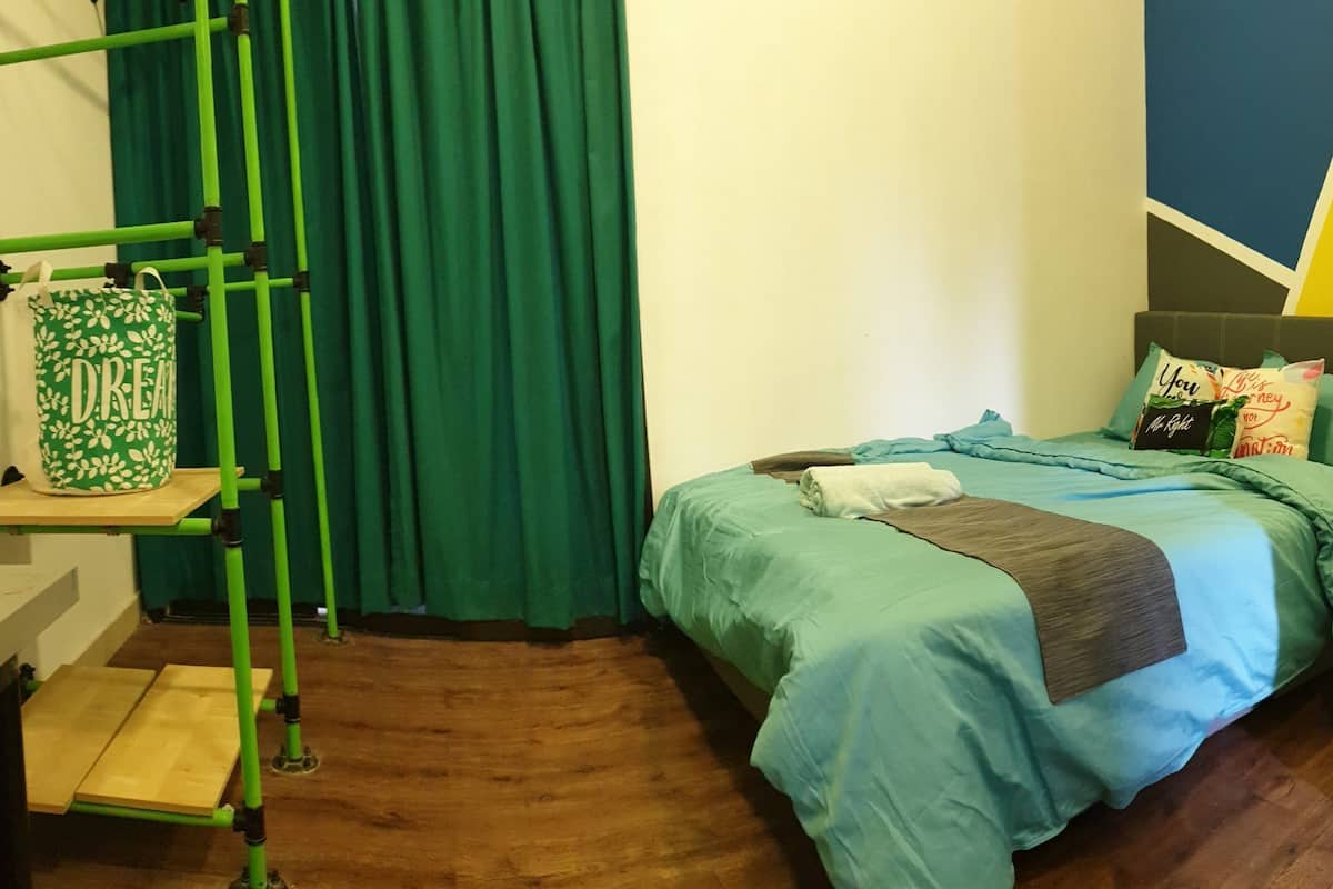 A bedroom with a bed, green curtains, a green and white patterned bag, and a wooden floor.