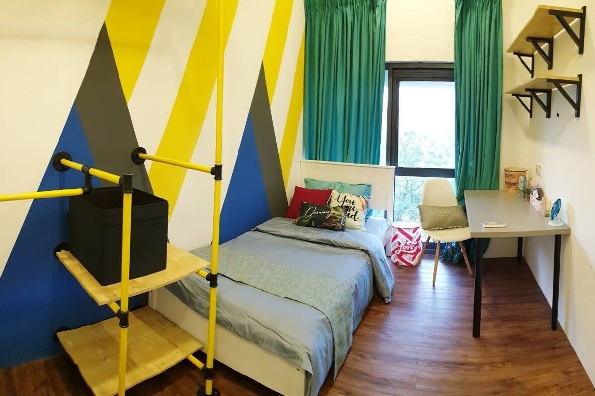 A compact bedroom with a bed, a desk, and a chair. There is a window with curtains, a shelf with items, and a wall with a geometric pattern.