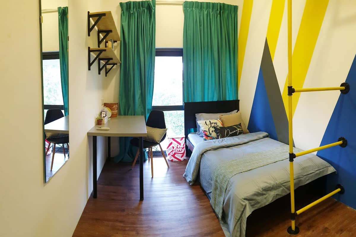 A compact room with a bed, a desk, and a chair. There is a window with curtains, a shelf, and a bed with pillows.