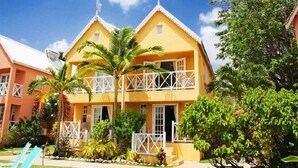 A yellow two-story building with a balcony, surrounded by tropical plants and a clear blue sky. - Villa located in St James on the west cost of Barbados with sea view and pool. (Fitts Village)
