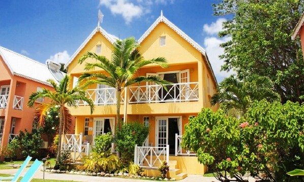 A yellow two-story building with a balcony, surrounded by tropical plants and a clear blue sky. - Villa located in St James on the west cost of Barbados with sea view and pool. (Fitts Village)