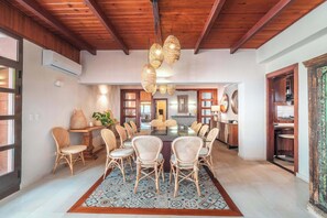 Un vaste espace dédié à la salle à manger, avec une grande table entourée de chaises en osier, un tapis à motifs et un plafond en bois.