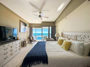 Habitación de hotel con cama, televisión, cómoda, silla, lámpara y vista al océano.