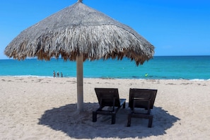 Una playa con una sombrilla de paja y dos sillas de madera.