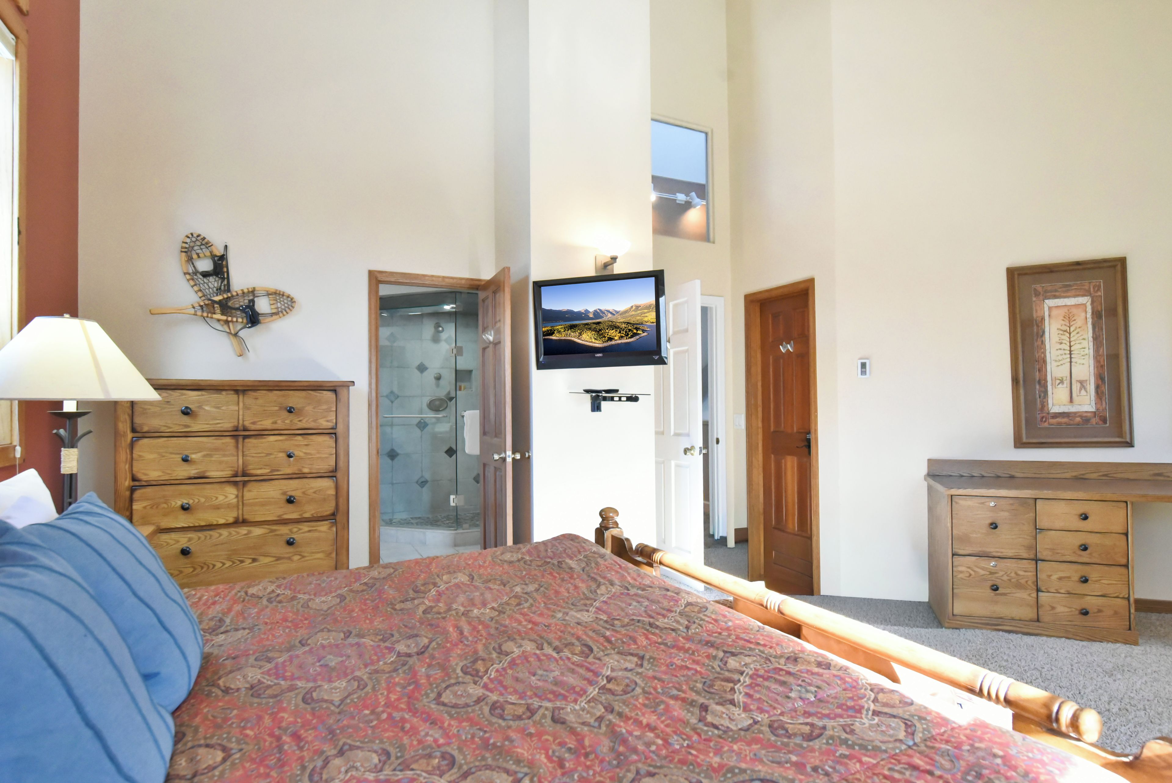 A bedroom with a bed, wooden dresser, a television mounted on the wall, and a door leading to another room.