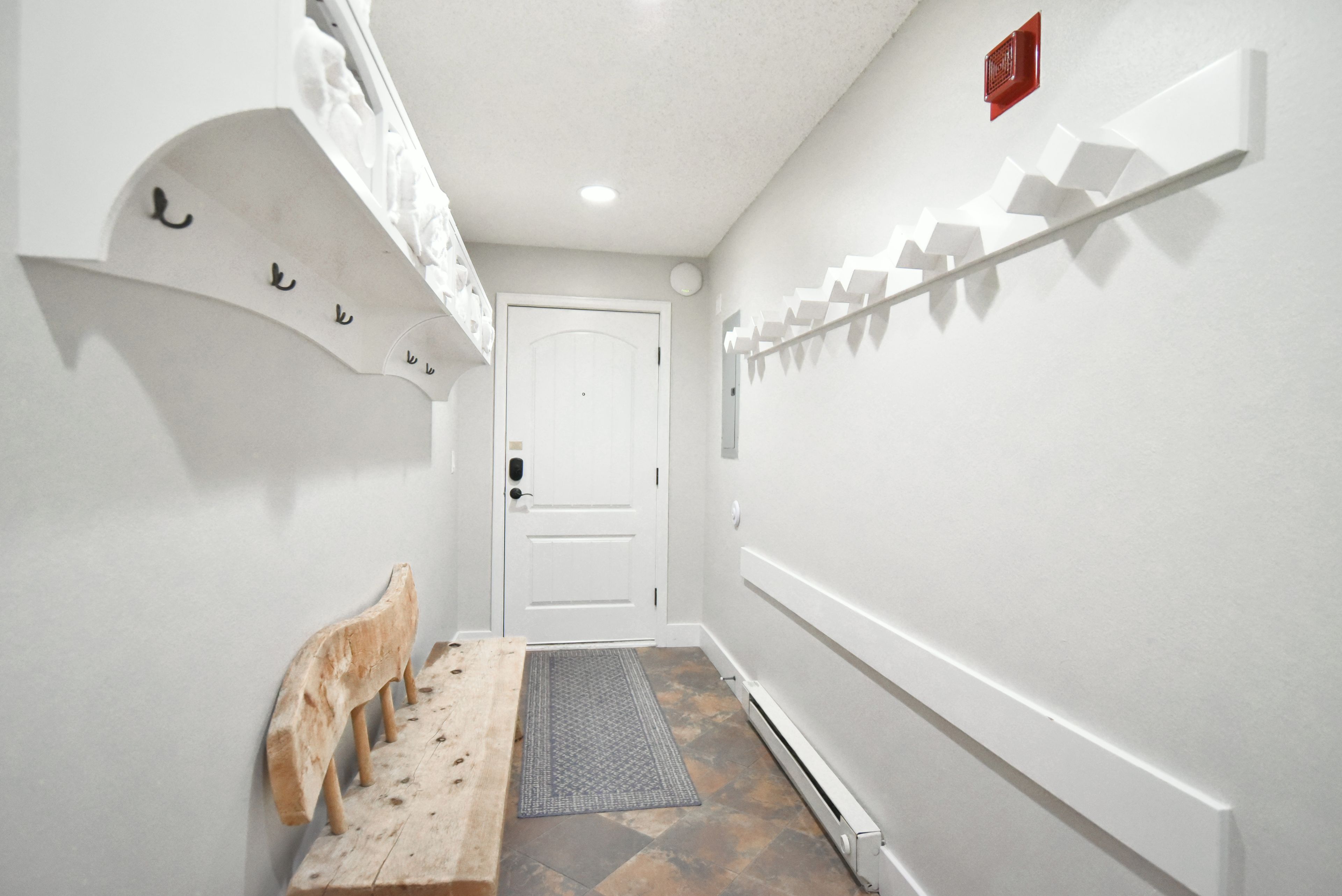 A narrow hallway with a white door at the end, a wooden bench, and a wall-mounted shelf with decorative items.