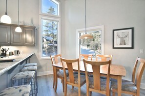 Une cuisine moderne avec des meubles en bois, un coin repas comprenant une table et des chaises en bois, et de grandes fenêtres offrant une vue sur les arbres.