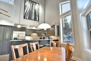 A modern kitchen with dark cabinets, a large island, pendant lighting, and a dining area with a wooden table and chairs.