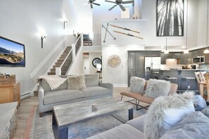 Un vaste espace de vie comprenant un canapé gris, une table basse et une cheminée. Un escalier mène à un niveau supérieur, et on aperçoit une cuisine au fond de la pièce.