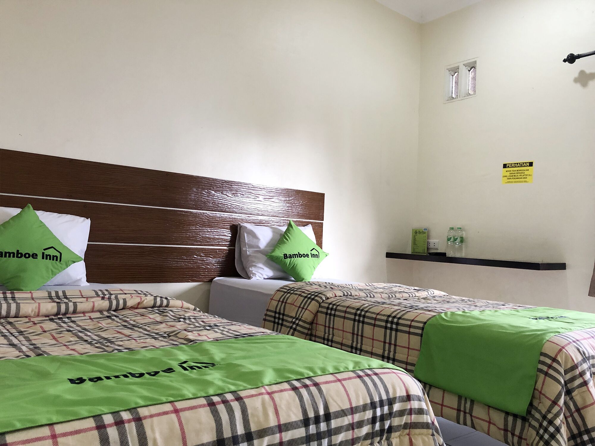 A hotel room with two beds, each with a plaid bedspread and green pillows with the hotel logo. There is a wooden headboard, a small shelf with toiletries, and a sign on the wall.