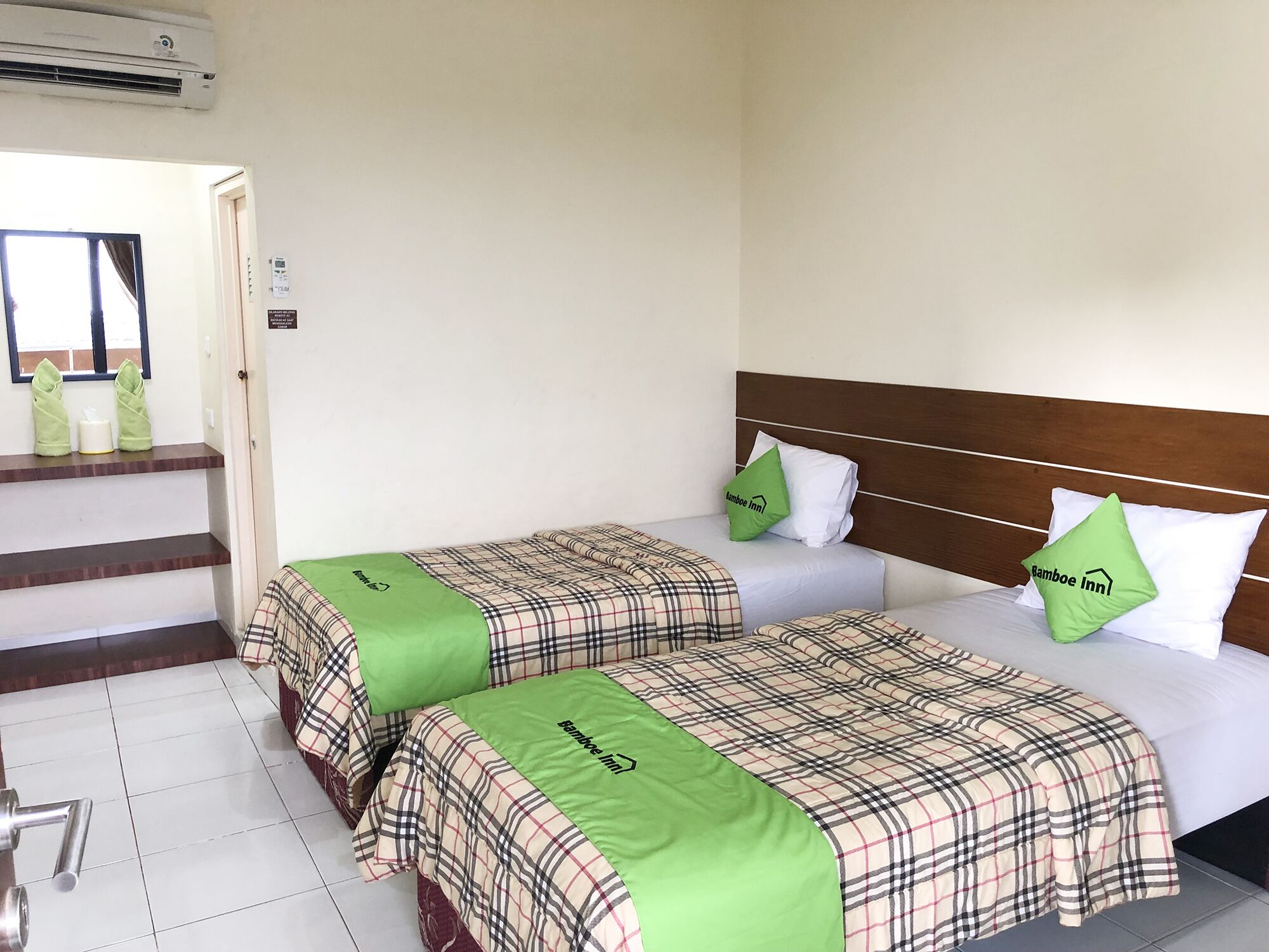 A hotel room with two beds, each with plaid bedding and green pillows, a wooden headboard, and a window with curtains.