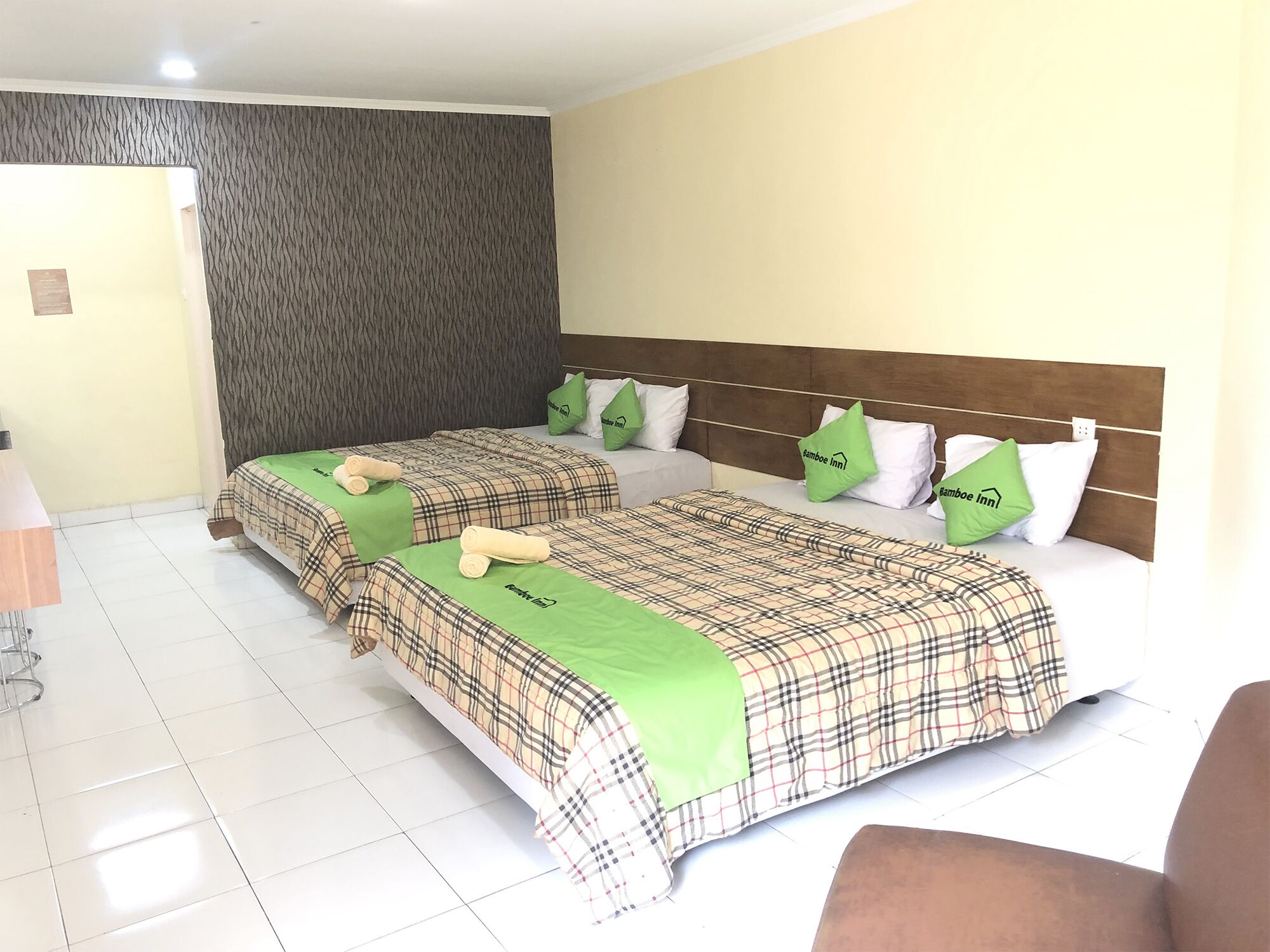 A hotel room with two beds, each with a plaid bedspread and green pillows, a wooden headboard, and a tiled floor.