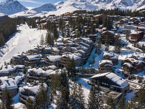 Un complejo turístico en la montaña cubierto de nieve con varias construcciones tipo chalet, pistas de esquí y densos bosques de pinos.