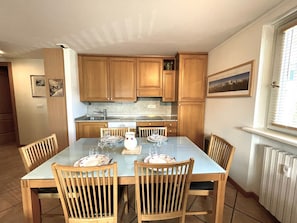 Un comedor con una mesa dispuesta para cuatro, sillas de madera, una cocina con armarios de madera, una ventana con persianas y un radiador.