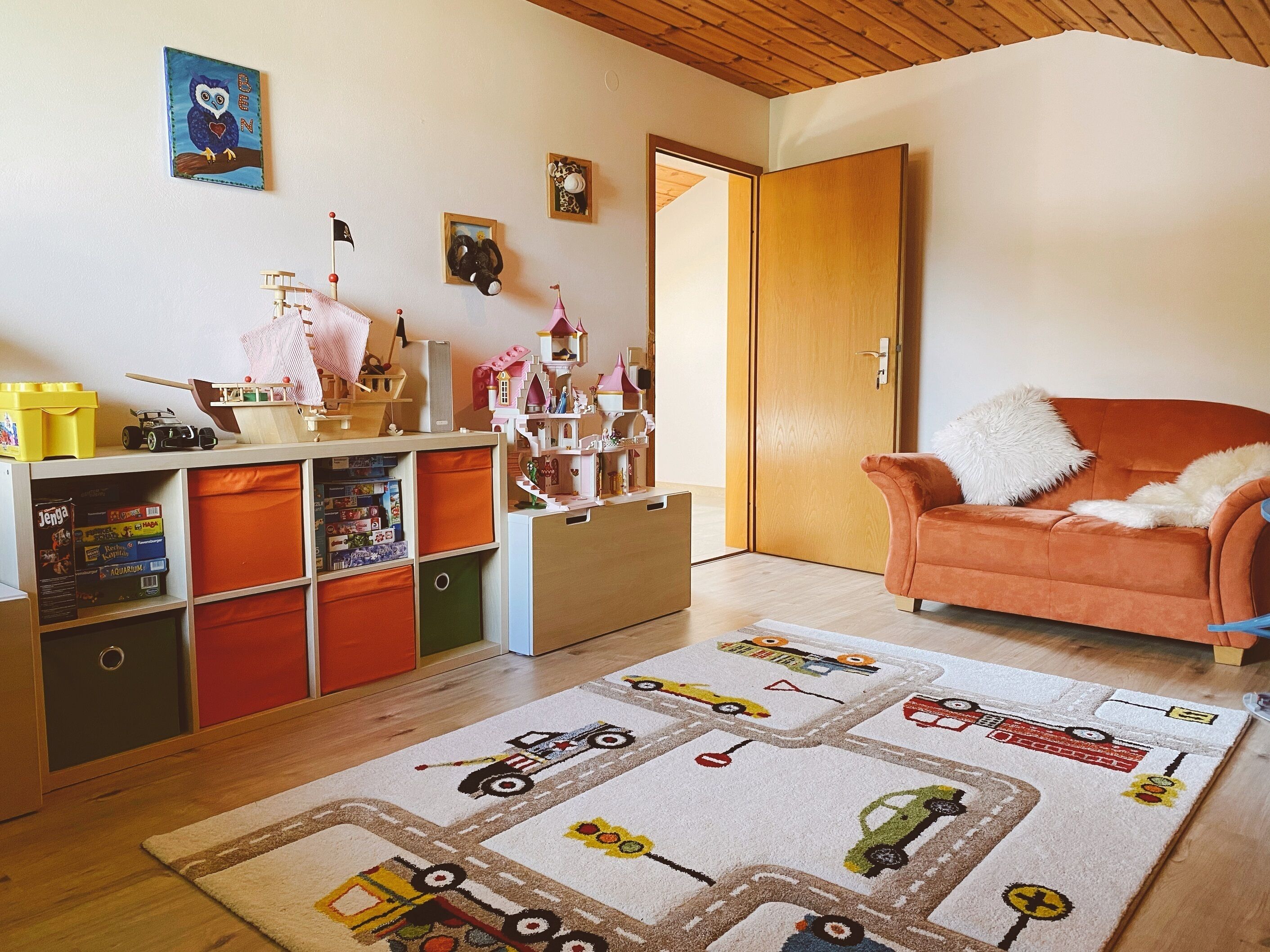 Un accogliente soggiorno con soffitto in legno, un divano arancione confortevole, un tappeto colorato a tema ferroviario e un’area giochi per bambini con giocattoli e contenitori.
