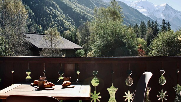 Mesa de comedor con pasteles y decoraciones colgantes, con vistas a un paisaje montañoso que incluye una casa y árboles.