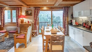 A cozy wooden dining room with a table set for four, a kitchen with white cabinets, and a balcony with a view of trees. - Spacious apartment in Chamonix - Sleeps 6 - Wifi and parking (Chamonix-Mont-Blanc)