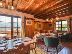 Une pièce chaleureuse avec un plafond en bois, un ensemble de canapés, une table à manger avec des chaises et une grande fenêtre donnant sur l’extérieur.
