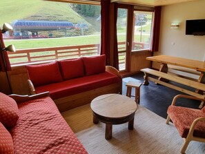 Una habitación con sofá rojo, mesa de madera y bancos, con vistas a un paisaje verde y un edificio al fondo.