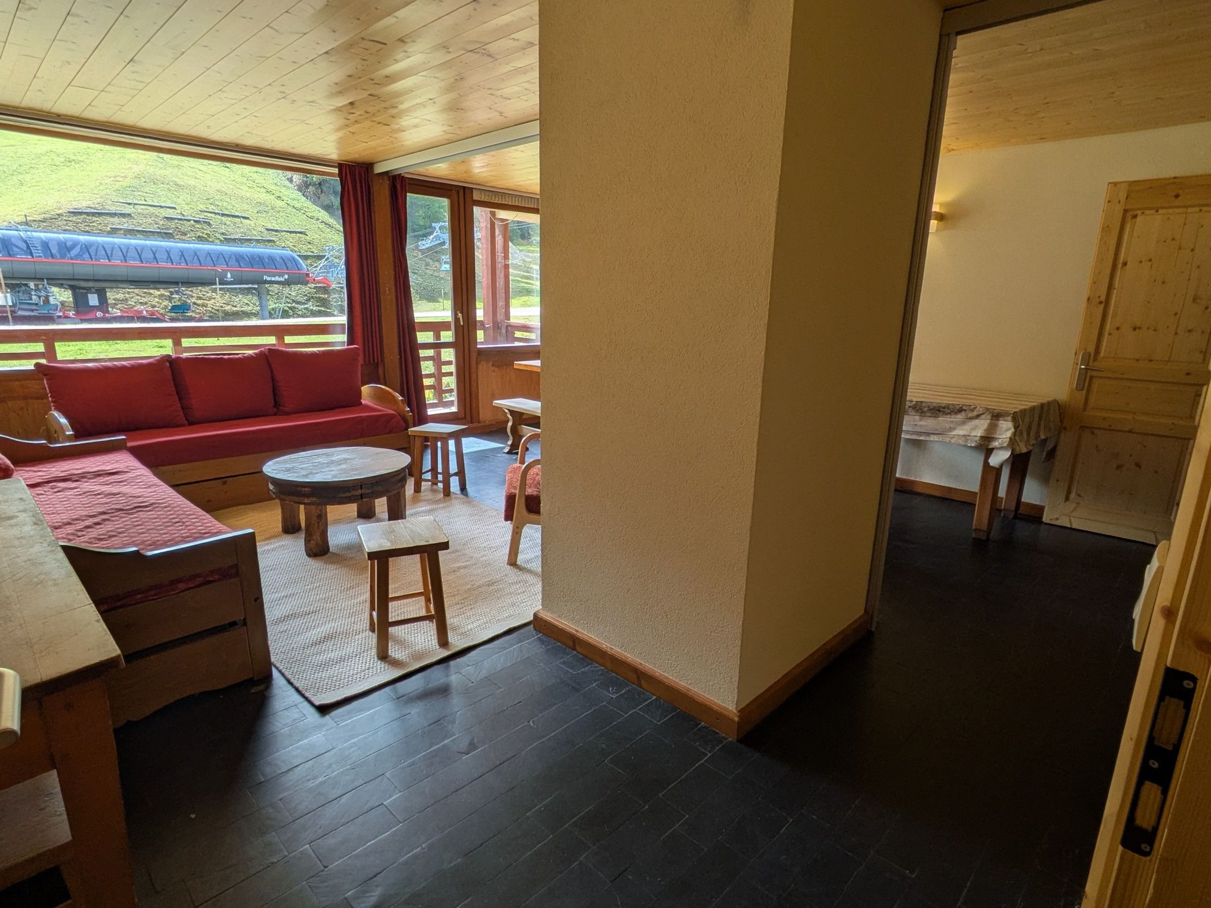 A room with a wooden ceiling, a sofa, a table, and chairs. There is a window with a view of a hill and a ski lift.