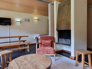 A cozy room with a wooden ceiling, a fireplace, a red upholstered chair, and a wooden table. - Large South Apt · Ski-in/out · Balcony · 8 pers. (La Plagne)