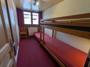 A small, single-room accommodation with a bunk bed, a small table, a radiator, and a window with curtains.