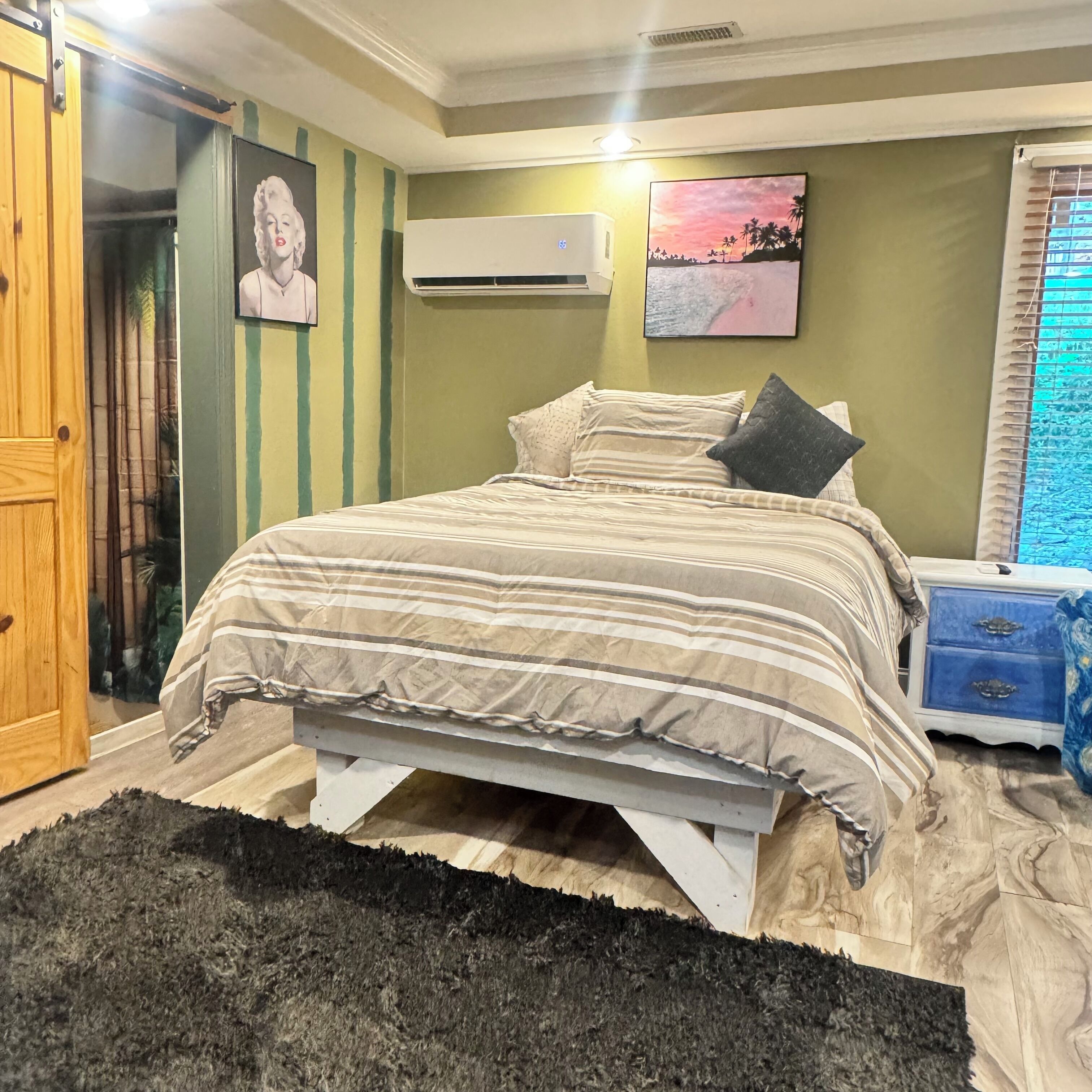 Een slaapkamer met een bed, een nachtkastje, een raam met zonwering en een wandairconditioner.