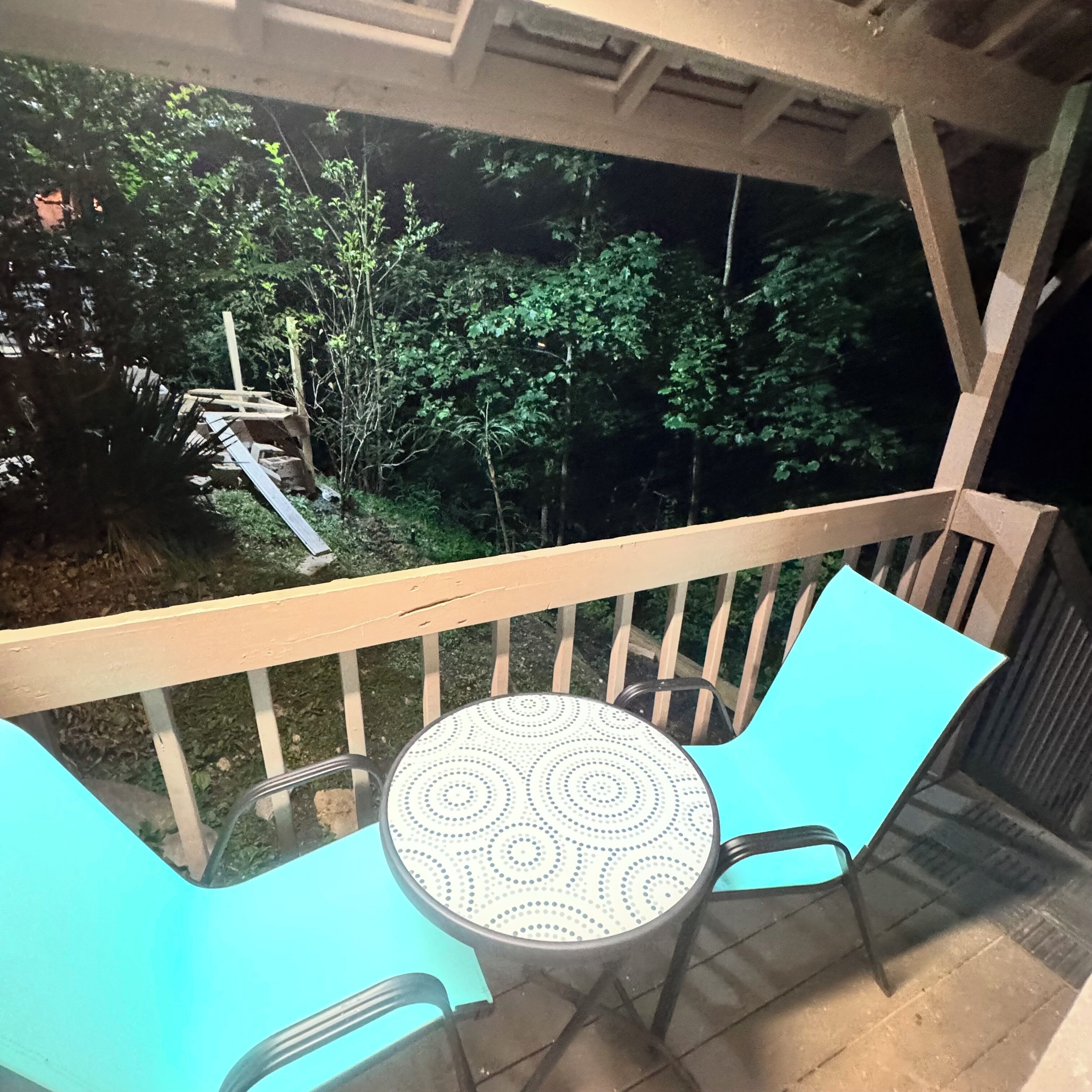 A balcony with a table and chairs, overlooking a garden with a wooden deck and trees.