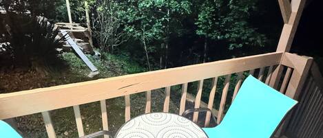 A balcony with a table and chairs, overlooking a garden with a wooden deck and trees.