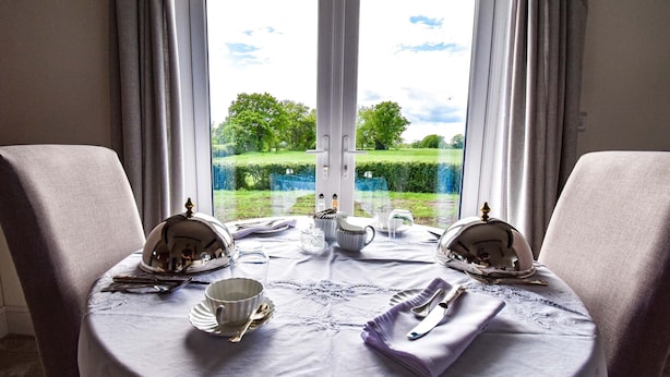 A dining table set for two with a view of a green landscape through a window. - The Cowhous at Abbey Green Farm (Whitchurch)