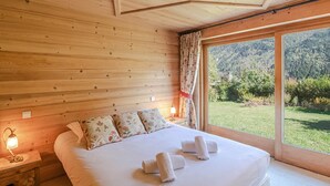 A wooden-paneled bedroom with a large bed, floral pillows, and a view of a green landscape through a large window. - Typical Chalet in Chamonix with Parking and Wifi, Near Center and Slopes (Chamonix-Mont-Blanc)