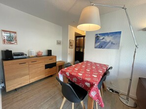 Een eethoek met een tafel bedekt met een rode, geruite tafelkleed, een houten kast met een speaker en een decoratief object, en een schilderij aan de muur.