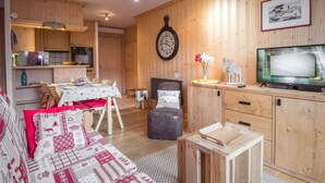 A cozy cabin-style room with a wooden interior, a dining table, a sofa with a red and white patterned cover, a television, and a clock on the wall. - Lovely apartment in Chamonix center, near slopes with parking (Chamonix-Mont-Blanc)