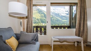 Une pièce avec un canapé gris, un petit banc blanc, une grande fenêtre donnant sur des montagnes enneigées et une lampe blanche.