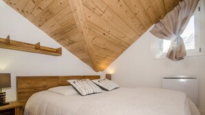 Une chambre avec un plafond en pente, une tête de lit en bois, un lit avec une couette blanche et des oreillers à rayures, une table de chevet avec une lampe et une fenêtre avec des rideaux.