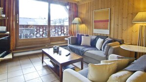 Un salon chaleureux avec un plafond en bois, une grande fenêtre avec des rideaux, un ensemble de canapés, une table basse et une lampe.