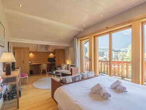 A bedroom with a large bed, a desk, a chair, and a view of the mountains through the windows.